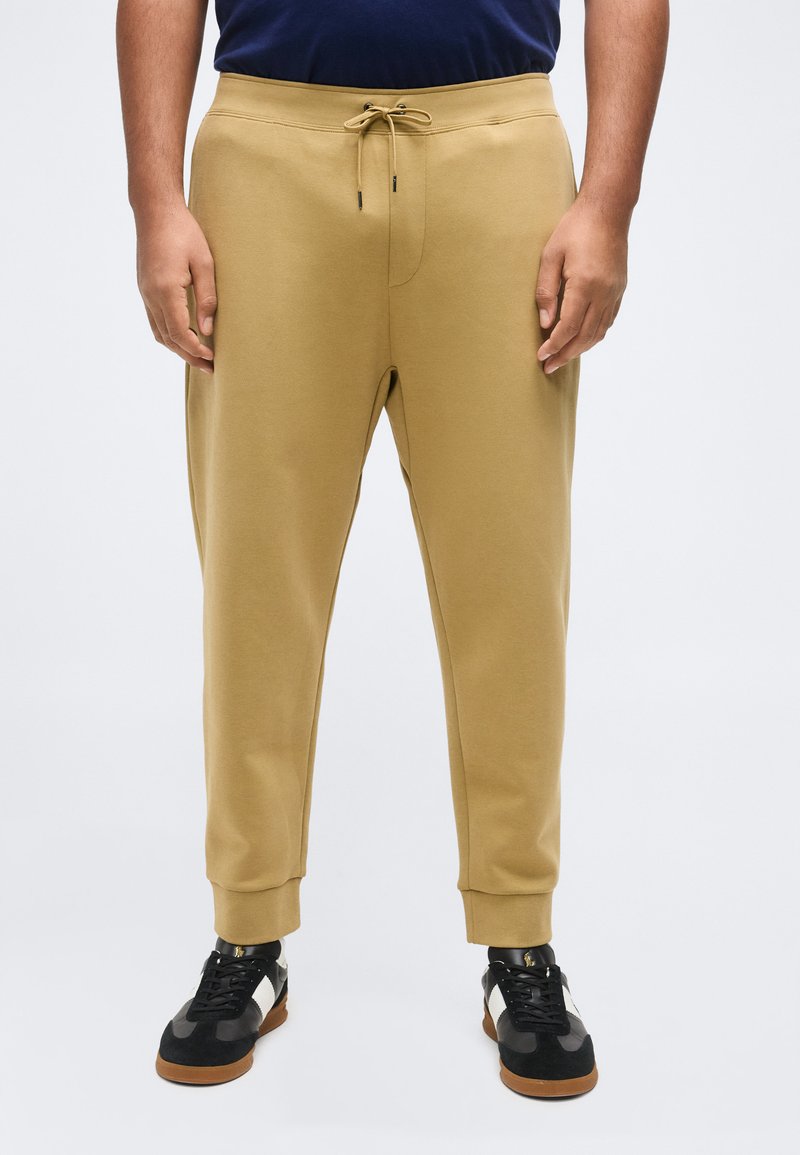 Ά νδρας που φοράει μπεζ παντελόνι jogger με κορδόνι στη μέση και μαύρα-άσπρα παπούτσια αθλητικά, μπροστά σε απλό φόντο.