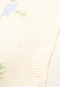 Κρεμ πλεκτή ζακέτα με λευκά κουμπιά και κεντητό μπλε λουλούδι με πράσινα φύλλα.