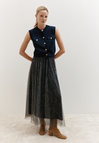 Haut sans manches bleu marine avec fermeture à bouton et deux poches poitrine, associé à une jupe maxi noire et beige à motif et des bottines beiges.