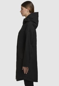 Veste à capuche noire au design épuré, avec une fermeture éclair et des poches latérales. Le matériau semble lisse et léger.