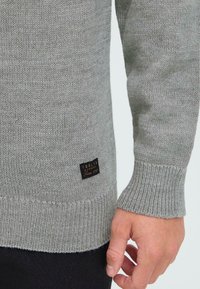 Grå strikkegenser med ribbede mansjetter og hem. Har en svart etikett på siden med tekst. Materialet har en myk, teksturert finish.