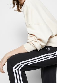 Krämfärgad sweatshirt med sänkta axlar, ihopparad med svarta sweatpants med tre vita ränder och en Adidas-logotyp på låret.