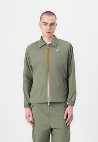 Jachetă ușoară de culoare verde măslinie cu guler ascuțit și fermoar frontal. Prezintă dungi galbene, albastre și roșii pe lângă fermoar.