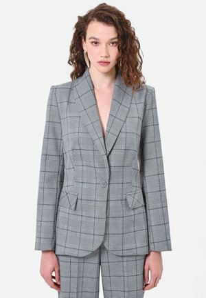Donna con capelli ricci che indossa una giacca di abito grigia a quadri e pantaloni abbinati, in piedi davanti a uno sfondo bianco neutro.