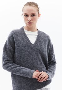Sweter z dekoltem w serek i długim rękawem w miękkim szarym materiale. Posiada luźny krój oraz ribbowe mankiety. Pod spodem widoczna jest biała koszulka z okrągłym dekoltem.