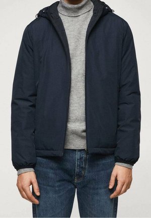 Mand iført en navy hættejakke over en grå ribstrikket rullekravetrøje og blå jeans, stående med armene afslappet ned langs siderne.