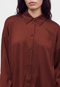 Femme portant une chemise marron ample à manches longues avec des boutons, à col pointu, montrée des épaules jusqu'en haut de la poitrine.