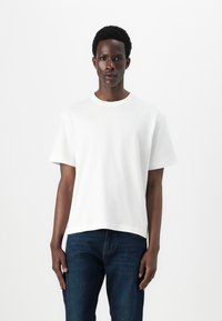 White crew neck t-shirt in cotton; short sleeves, relaxed fit, straight hem. Worn with dark blue denim jeans, minimal design, no visible logos.