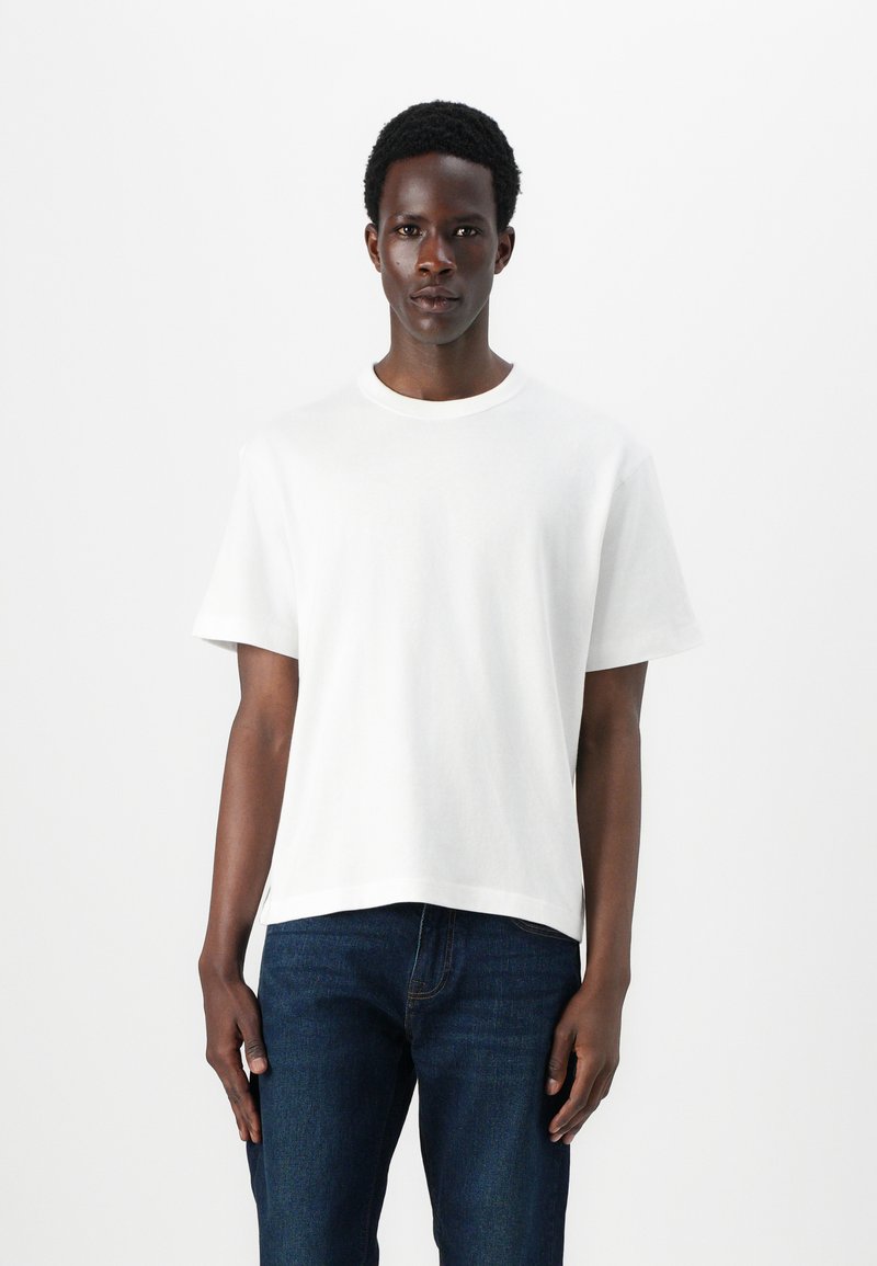 White crew neck t-shirt in cotton; short sleeves, relaxed fit, straight hem. Worn with dark blue denim jeans, minimal design, no visible logos.