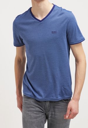 Mand iført en blå T-shirt med korte ærmer og V-udskæring med diskrete striber og grå jeans, stående med den ene hånd nær lommen.