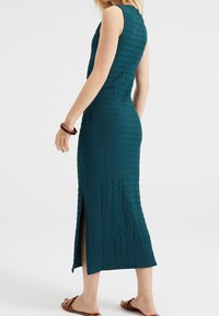 Ärmlös turkossfärgad midi-klänning med struktur och slits på sidan, båren av en person, parat med bruna sandaler och matchande armband på ena handleden.