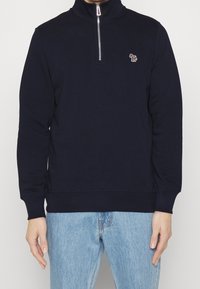 Felpa navy a maniche lunghe con colletto alto, zip frontale a metà e un piccolo logo ricamato sul petto; realizzata in tessuto morbido.