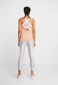 Débardeur de sport rose clair avec un design dos croisé et une découpe ouverte ; porté avec des leggings argentés brillants et des baskets légères.