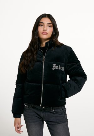Жена с дълга тъмна коса, облечена в черно пухено яке с цип и "Juicy" от камъчета на гърдите, комбинирана със тъмни джинси.