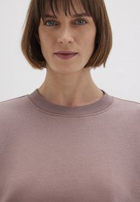 Light mauve sweatshirt with a ribbed crew neckline and a soft, textured fabric. Plain design without patterns or embellishments.