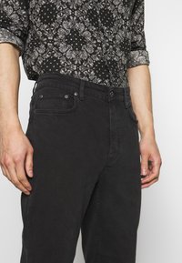 Černé džíny z denimového materiálu s hladkou texturou, pěti kapsami a poutky na opasek, doplněné černobílou vzorovanou košilí.