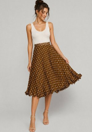 Femme aux cheveux en chignon portant un top blanc sans manches et une jupe midi plissée marron à pois blancs, tenant le bord de la jupe, sandales à talons beige.