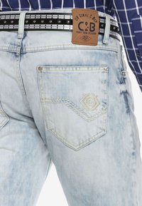 Helleblaue Denim-Jeans mit einem verwaschenen Finish, ausgestattet mit genähten Gesäßtaschen und einem braunen Lederetikett. Schwarzer Nietenimitat-Gürtel enthalten.