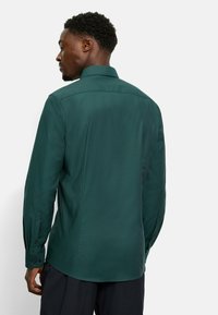 Camicia a maniche lunghe di un verde scuro con un tessuto texturizzato, polsini abbottonati e colletto classico. Presenta cuciture sottili e un orlo curvato.