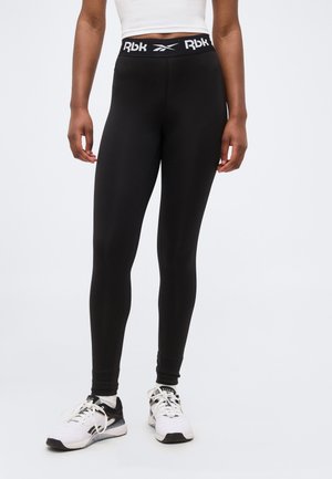 Femme portant un legging noir Reebok avec une taille marquée du logo, des baskets blanches et un top court blanc sur un fond clair uni.