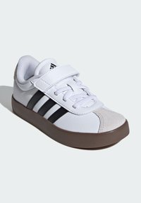 Biały sneaker z czarnymi paskami, brązową podeszwą, paskiem na rzep i białymi sznurówkami na jasnym tle, ustawiony pod kątem, ukazujący zewnętrzną stronę i przód.