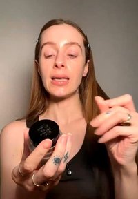 Femme aux longs cheveux tenant et montrant un petit pot de produit de soin de la peau ou cosmétique sur un fond uni.