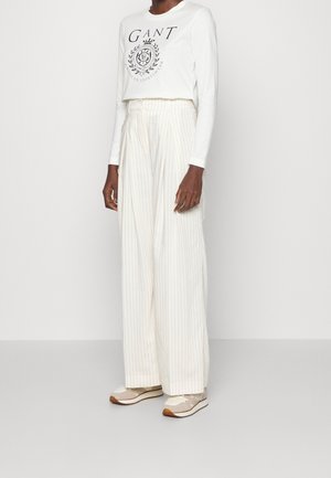 Chemise blanche à manches longues avec un logo imprimé, associée à un pantalon crème à rayures verticales et jambes larges, ainsi qu'à des baskets de couleur claire.