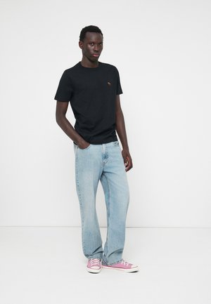 Noor mees seisab käed taskus, seljas must t-särk, helesinised teksad ja roosad spordijalatsid, taustal on lihtne valge taust.