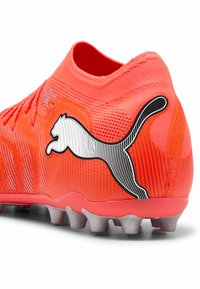 Punainen jalkapallokengän päällinen on teksturoitua verkkoa, jossa on näkyvä mustavalkoinen Puma-logo ja joustava pohja, jossa on useita valkoisia nappuloita.