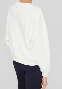 Sudadera blanca de gran tamaño con hombros caídos, puños y dobladillo acanalados, y un tejido suave y texturizado. Llevada con pantalones oscuros.