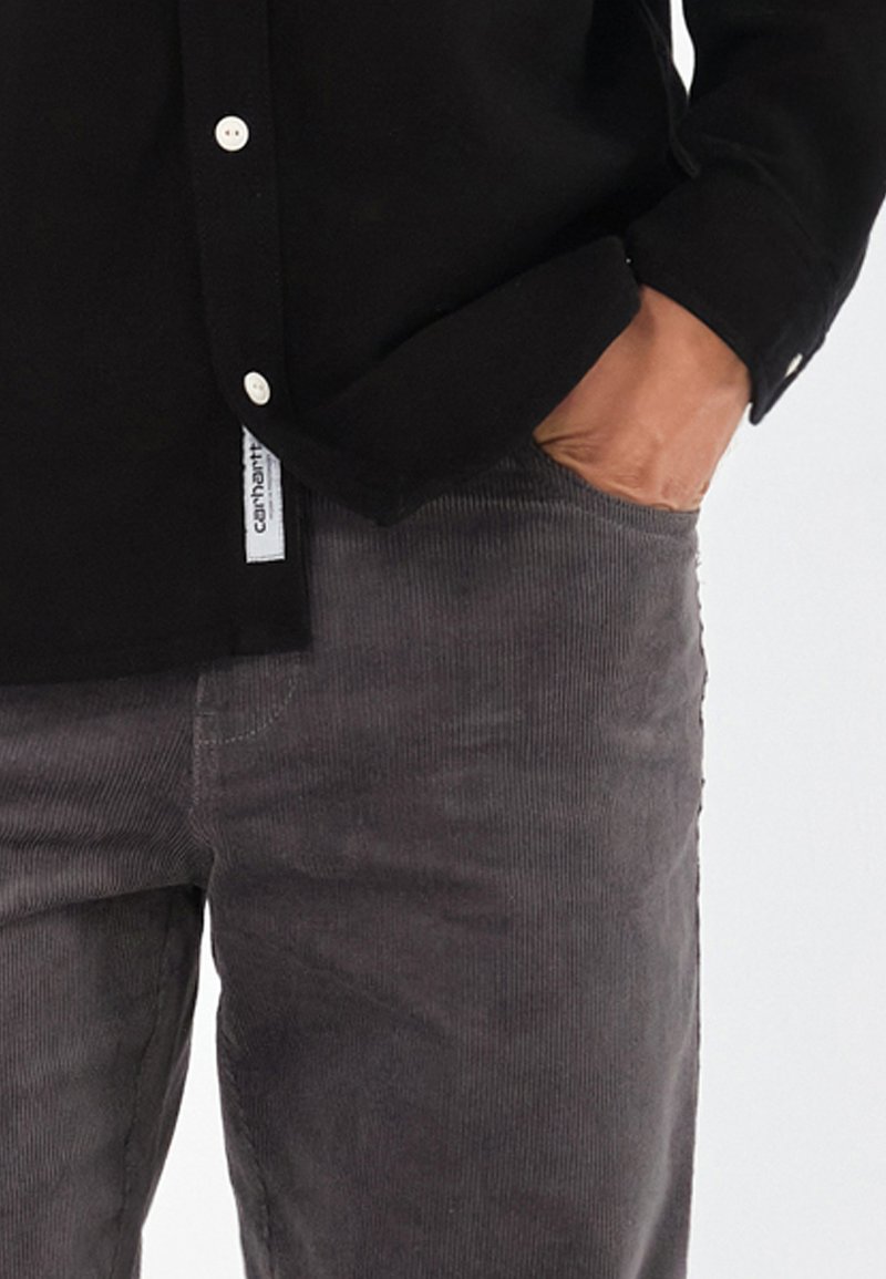 Camisa negra de botones con botones blancos, combinada con pantalones de pana grises que tienen un bolsillo lateral y un tejido texturizado.