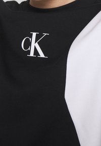 Camiseta de algodón negra con un panel diagonal blanco, escote redondo y un logo "cK" blanco en el pecho. Textura suave.