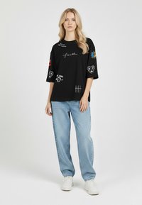 Black oversized t-shirt with various colorful embroidered graphics and text on sleeves. Paired with light blue straight-leg jeans and white sneakers.