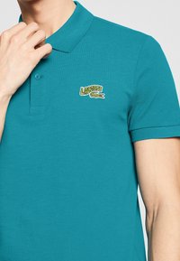 Teal pikétröja i texturerat tyg, med krage, två knappar och en broderad Lacoste-logotyp i grönt och rött på bröstet.