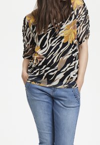 Blusa floreale nera e gialla con maniche corte, caratterizzata da un design fluido, abbinata a jeans blu chiaro con cuciture decorative.