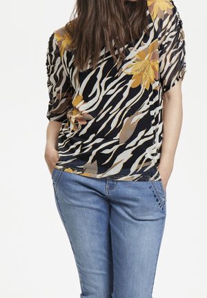 Blusa floreale nera e gialla con maniche corte, caratterizzata da un design fluido, abbinata a jeans blu chiaro con cuciture decorative.