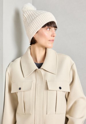 Bonnet en tricot crème avec un motif texturé et un pompon poilu. Le modèle porte une veste crème avec deux poches poitrine et un large col.