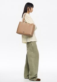 Torebka tote w kolorze tan, o sztywnej konstrukcji, z podwójnymi uchwytami u góry i wytłoczonym logo. Połączona z koszulą w paski z długim rękawem i spodniami z szerokimi nogawkami.