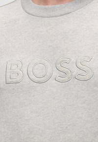 Sweat-shirt gris clair avec des lettres brodées en relief formant le mot « BOSS » sur la poitrine, près de l'encolure.