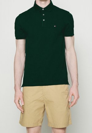 Ciemnozielona koszulka polo z kołnierzykiem, listwą guzikową i małym logo na piersi; zestawiona z krótkimi spodniami w kolorze khaki. Gładka tekstura materiału.