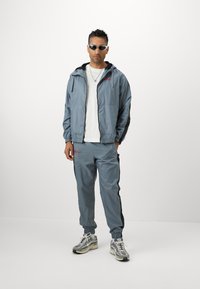Grå nylon træningsdragt med sorte sidelister, der har en hættetrøje og elastiske manchetter. Grå sneakers med et mønsterdesign.