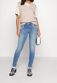 Beige stickad väst över en vit t-shirt, ihop med ljusblå skinny jeans med svängda fickor och skor med metalliska detaljer.