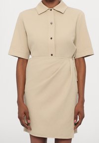 Femme portant une robe-chemise beige à manches courtes avec col, boutons devant et une ceinture à nouer à la taille style portefeuille, sur fond blanc uni.