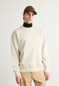 Beige särk ümmarguse kaelusega ja varrukate äärised, millel on rinna peal diskreetne logo. Kantud musta kõrge kaelusega särke peal.