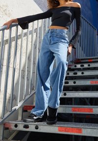 Světle modré džínové kalhoty s vysokým pasem a volným střihem, doplněné černým topem s odhalenými rameny a černými teniskami, na kovovém schodišti.