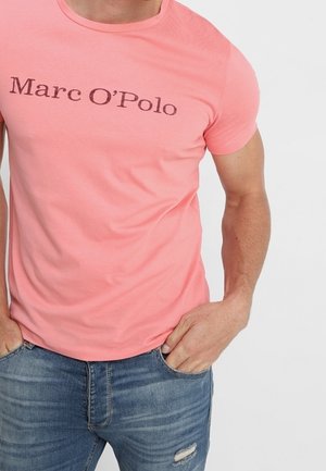 Mies pukeutuneena vaaleanpunaiseen Marc O'Polon t-paitaan ja sinisiin repeäviin farkkuihin, kädet taskuissa, yksiväristä valkoista taustaa vasten.