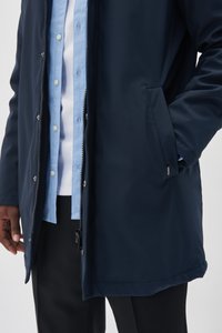 Chaqueta impermeable azul marino con cremallera frontal, cierres a presión, bolsillos laterales y textura suave; combinada con una camisa de botones azul clara.