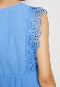 Haut bleu avec des détails en dentelle délicate sur les manches, un col rond et une taille froncée pour une silhouette féminine.