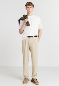 Pantaloni beige a pieghe, t-shirt bianca, giacca oliva appoggiata sulla spalla, cintura nera e scarpe slip-on marroni. Texture dei tessuti lisci.