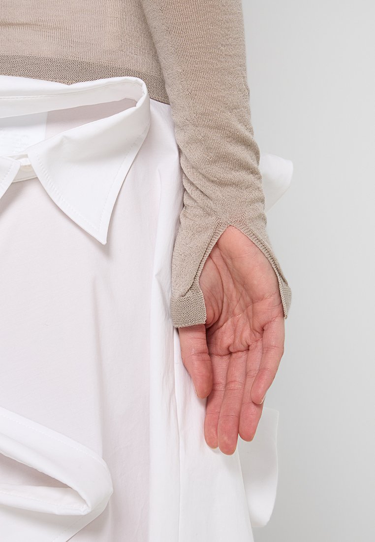 Beige enganliegendes Langarm-Oberteil mit Daumenlöchern, kombiniert mit einem weißen Kleidungsstück mit geschichteter Optik und sichtbarem Kragen. Die Hand ist entspannt.